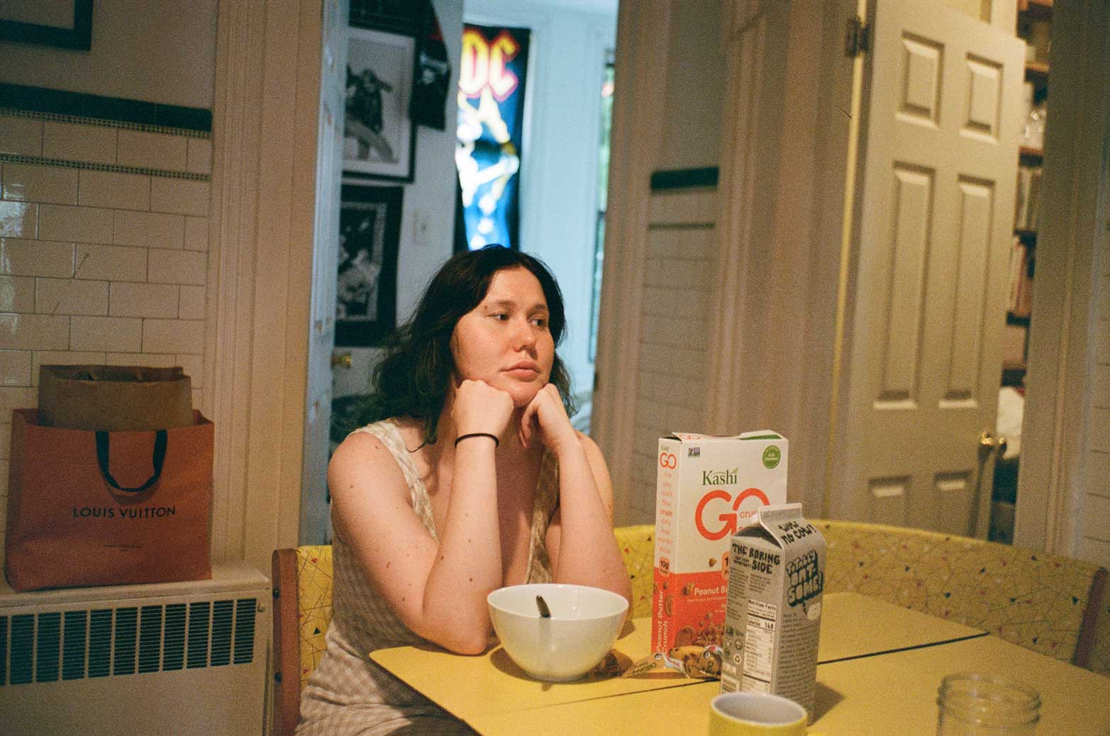 A light skinned person sits with their chin resting on their hands, looking to the right of the image, as if deep in thought. They sit at a yellow kitchen table with a bowl of cereal. In the background we can see an AC/DC poster in the window of another room.