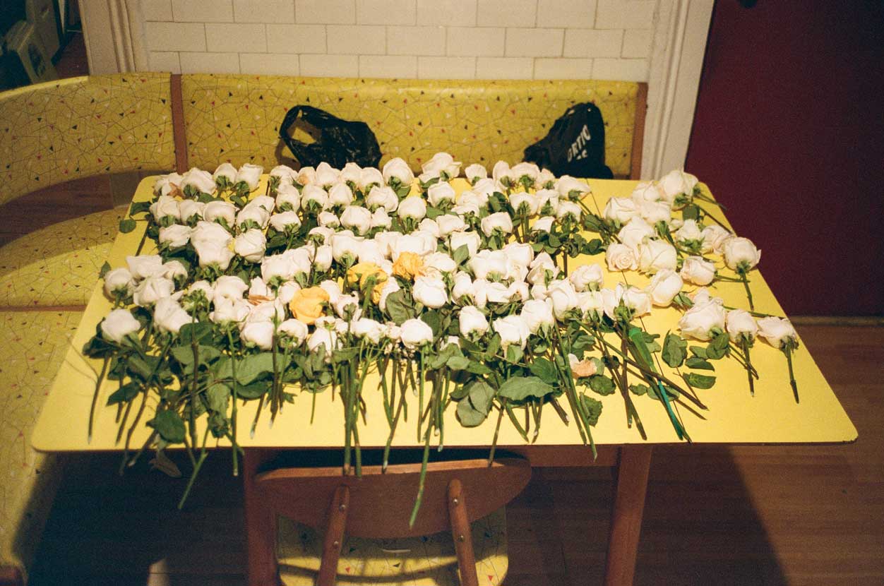 An analogue photograph of a yellow kitchen table covered with a hundred or so white roses with green stems. There is a yellow patterned bench that wraps around the table, on the bench are two black plastic bags, obscured by the table. There is one chair in front of the photographer tucked under the table.