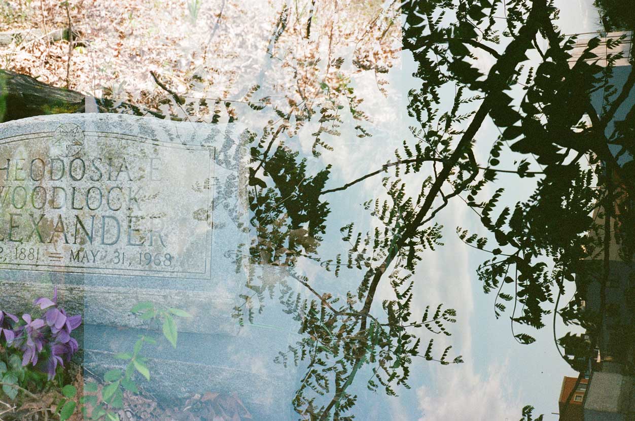 A double-exposed analogue photograph. Two images layered on top of each other. In one image a gravestone has text below a pentagram reads: HEODOSIA E., WOODLOCK, EXANDER, 1881 - MAY 31 1968. There are purple flowers at the bottom of the stone. The second photograph is pointed directly up to the sky and the silhouette of a tree covers the image, the tops of buildings can be seen along the right hand side of the frame.