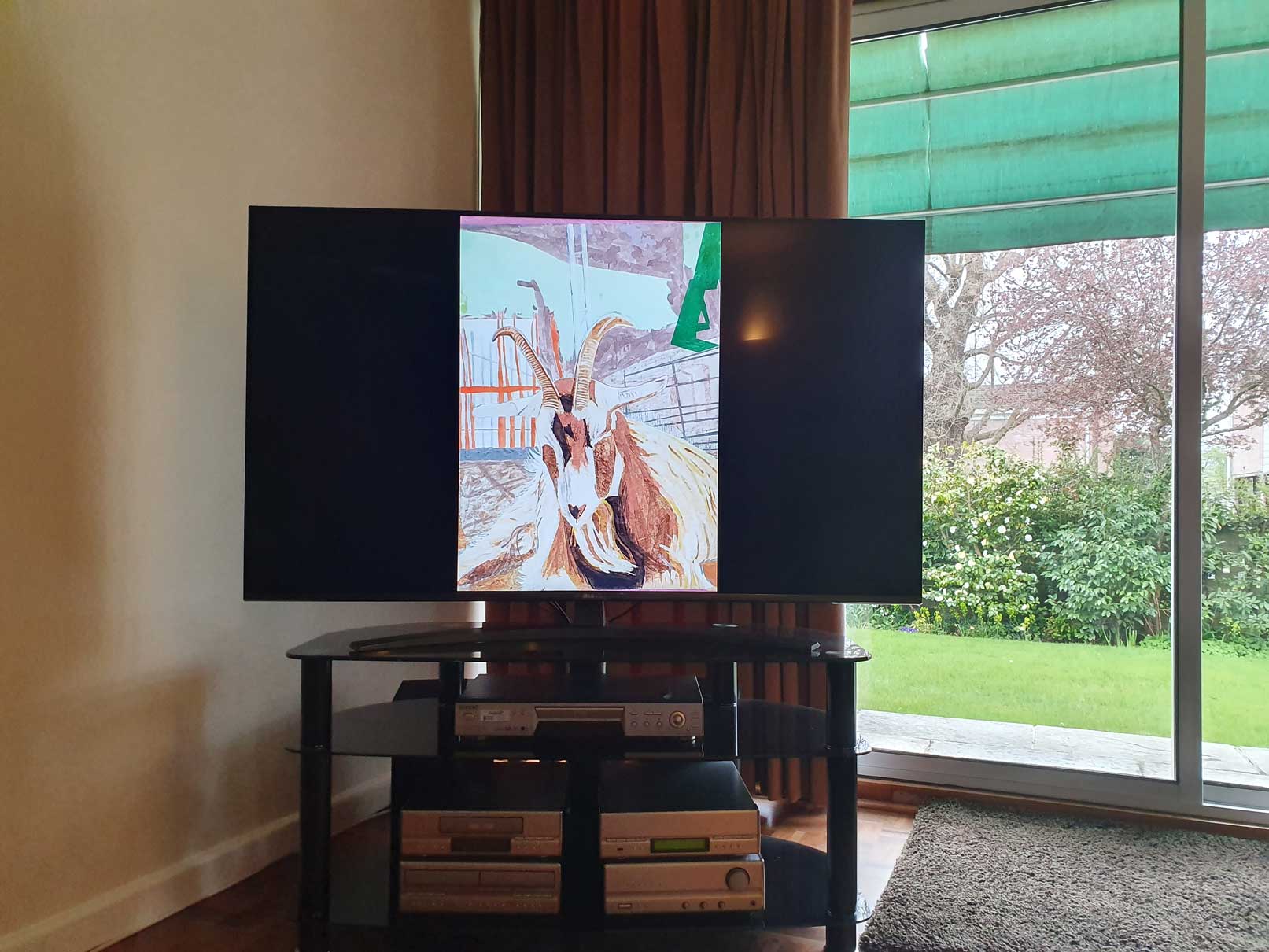 An image of a painting by the artist Alice Mendelowitz is displayed on a TV in the corner of a room, next to a glass sliding door that leads to a garden.