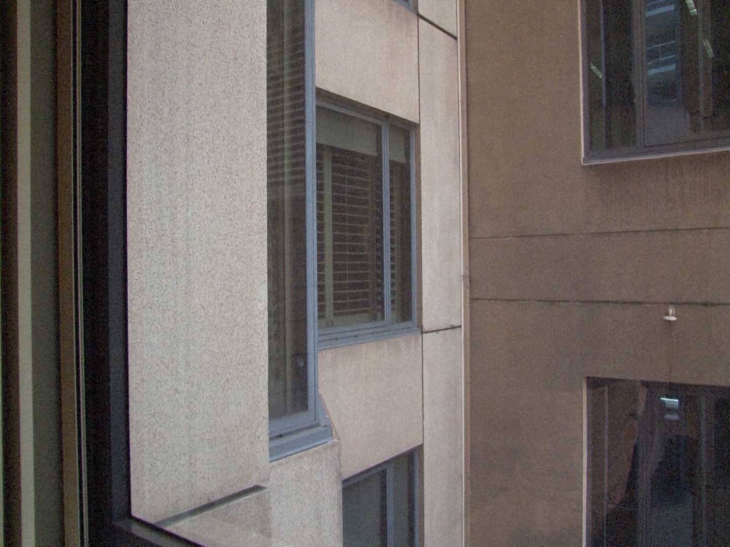 A film still, looking out of a window into a shady concrete courtyard. Other windows also look into the courtyard, some with blinds drawn down over the windows. We cannot see into the windows.