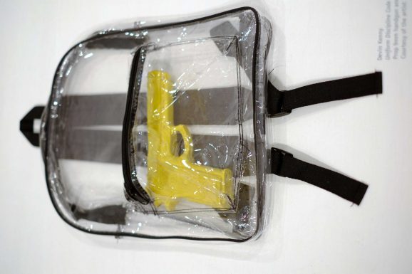 a see-through backpack on a white background with black zippers. A yellow plastic gun inside the back pocket.